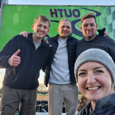 Four people smiling for a selfie in front of a green sign, outdoors on a sunny day.