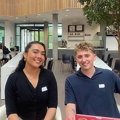 A young woman and man smiling in a university cafe setting, both wearing name tags.