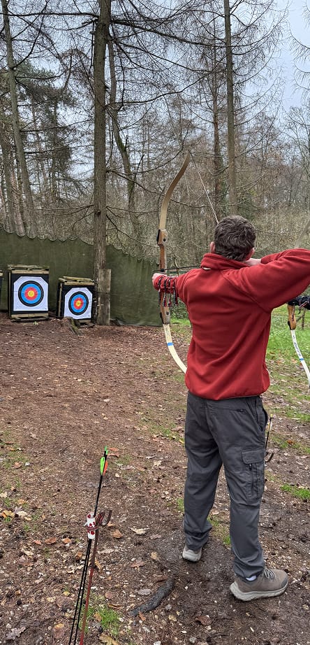 A person in a red fleece standing in a forest, aiming a recurve bow at two archery targets.