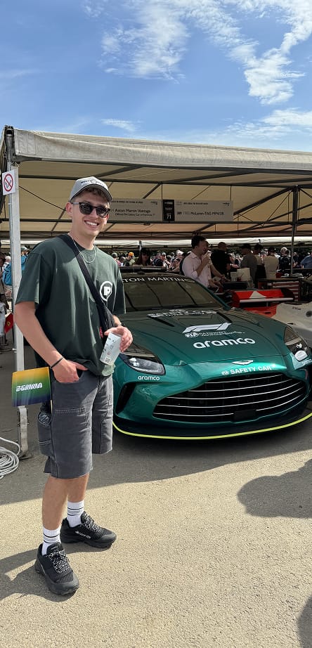 A smiling man in a green shirt stands in front of a green Aston Martin Formula 1 safety car at a trackside display.