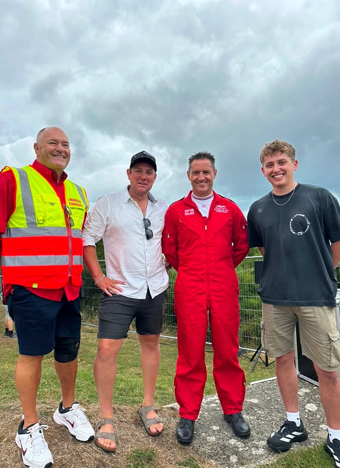 Four men standing outside in front of a chain-link fence, one wearing a high-visibility event jacket and another in a red flight suit.