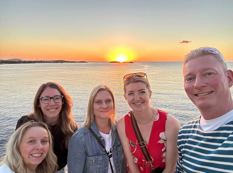 A group of five colleagues posing for a selfie during a scenic ocean sunset.