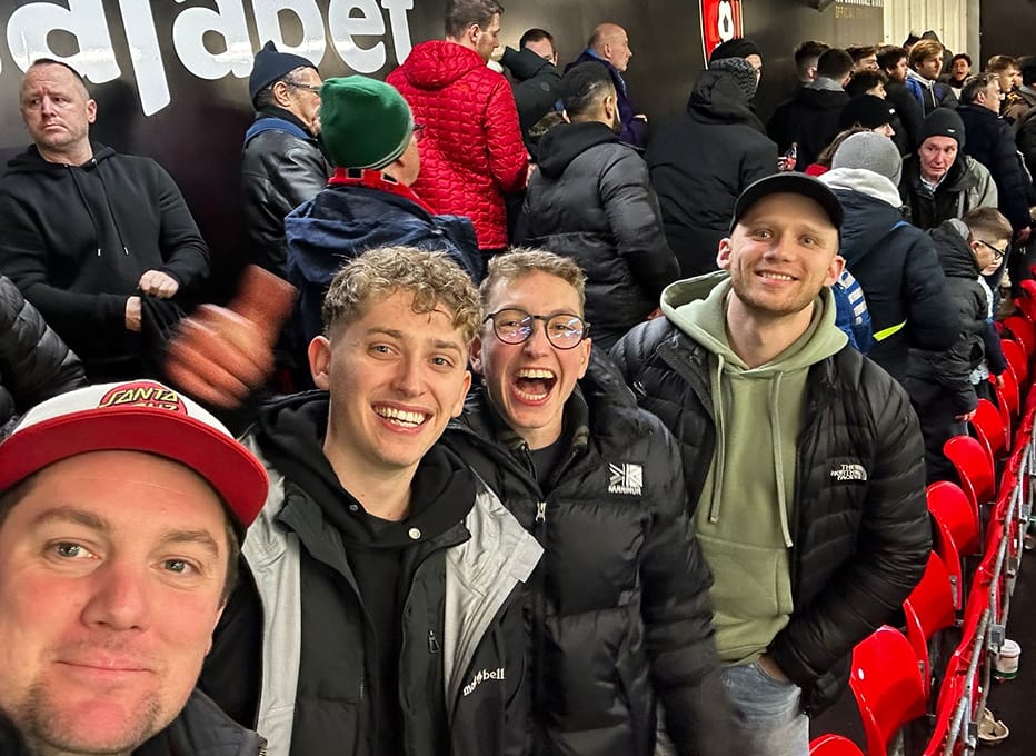 Three smiling young men taking a selfie in the stands at a crowded stadium with red seats.