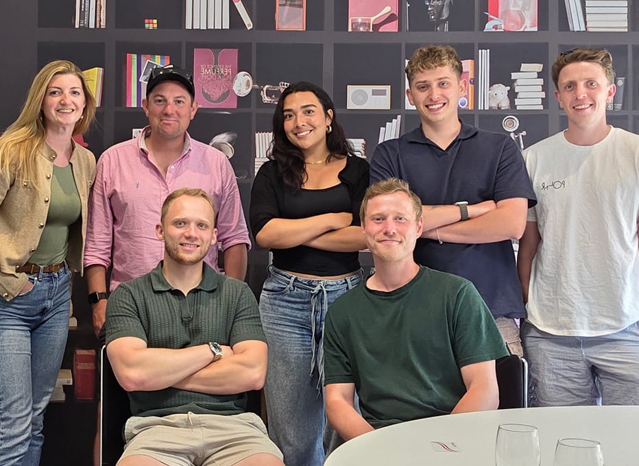 A group of seven colleagues smiling for a photo in front of a decorative wall with bookshelf motifs.